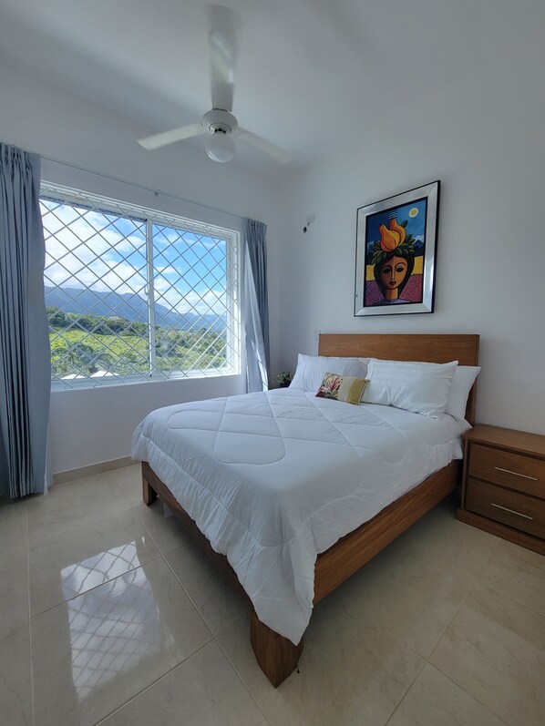 A bedroom with a bed, a ceiling fan, a window with curtains, a nightstand, and a framed picture on the wall.