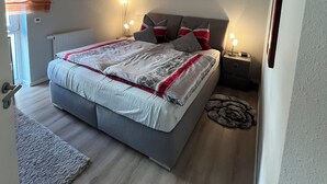 Un dormitorio con cama, mesitas de noche, radiador, una obra de arte en la pared y una ventana con cortinas.