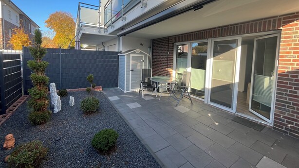 Un edificio residencial moderno con un patio que cuenta con una mesa y sillas, un cobertizo pequeño y un jardín vallado con rocas decorativas y arbustos.