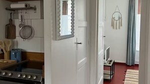 A kitchen with a stove, oven, and hanging utensils. There is a door with a mirror and a hallway leading to a room with a TV and a window with curtains. - The Pearl of the Cliffs (Le Tréport)