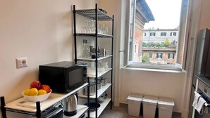 A compact kitchen with a microwave, shelves with glassware, a bowl of fruit, and a window with a view of buildings. - Beautiful house in the heart of Rome 20meters from Piazza Venezia/Trevi Fountain (Roma)
