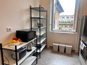 A compact kitchen with a microwave, shelves with glassware, a bowl of fruit, and a window with a view of buildings. - Beautiful house in the heart of Rome 20meters from Piazza Venezia/Trevi Fountain (Roma)