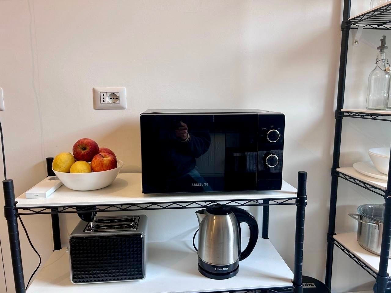 Cozinha equipada com micro-ondas, chaleira e uma tigela de frutas numa prateleira.