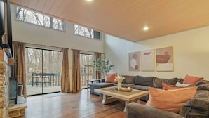 A spacious living room with a large sofa, a coffee table, and a fireplace. There are large windows and a sliding glass door leading to an outdoor area.