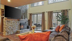 A cozy living room with a stone fireplace, a large flat-screen TV, a comfortable sofa, and a dining area visible through glass doors.