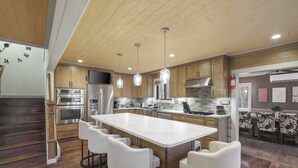 A modern kitchen with wooden cabinets, stainless steel appliances, a large island with bar stools, pendant lighting, and a visible clock on the wall.