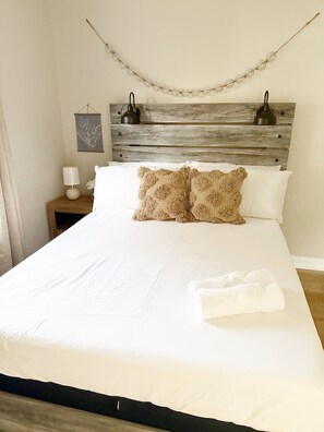 A neatly made bed with white linens and two decorative pillows, a wooden headboard, a bedside table with a lamp, and a wall-mounted light fixture. - Spring Street Suite D (Evansville)
