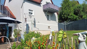 Une maison à deux étages, avec un toit recouvert de tuiles rouges, un balcon et un jardin agrémenté de diverses plantes et entouré d’une clôture.