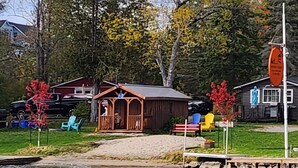 Una cabaña junto al lago con un muelle de madera, un pequeño porche y sillas Adirondack de colores.