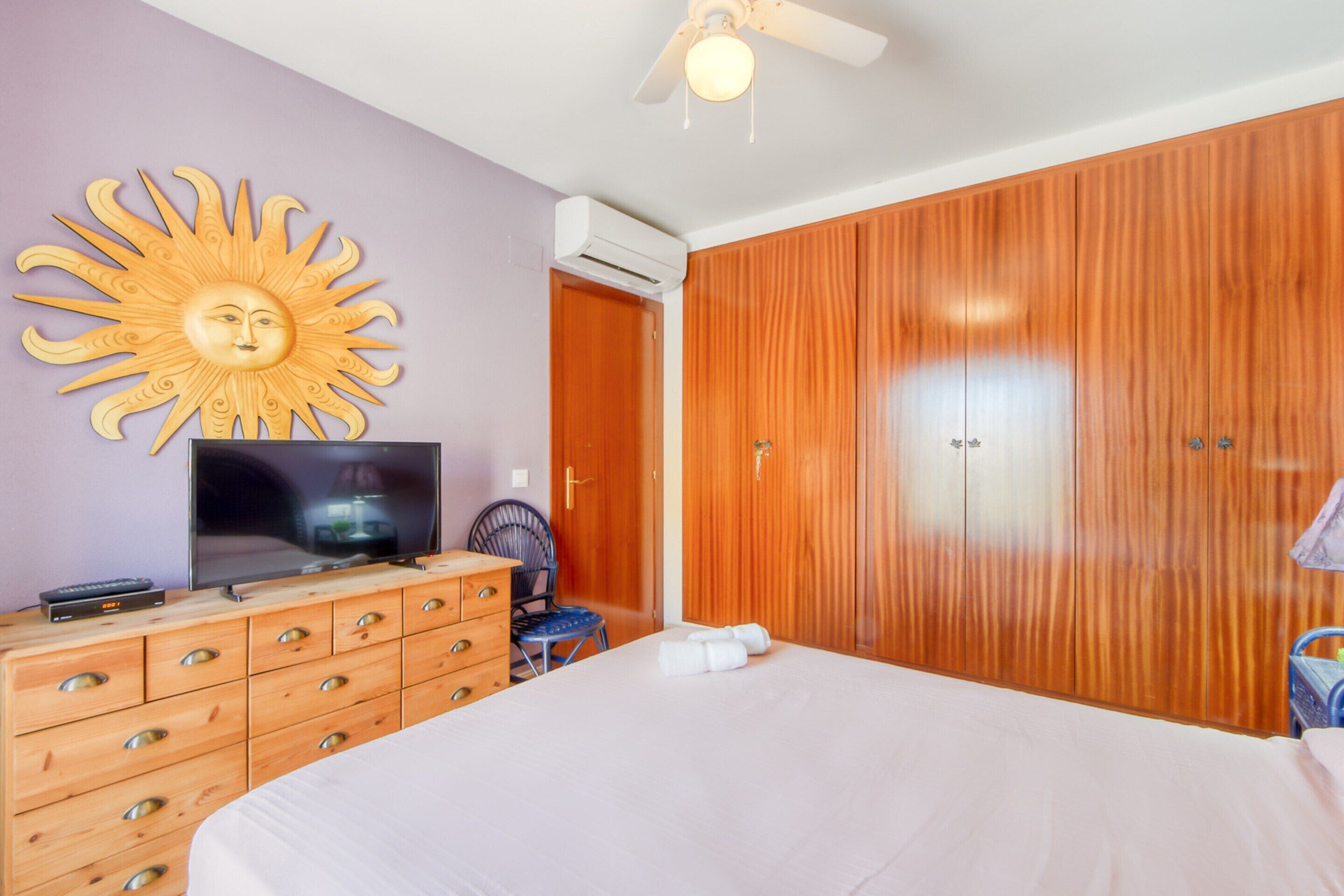 Une chambre à coucher comprenant un lit, une armoire en bois, un téléviseur posé sur une commode, une décoration murale en forme de soleil et un ventilateur de plafond.