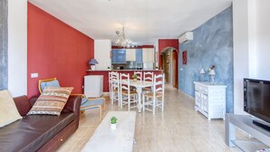 A living room with a sofa, coffee table, and TV, a dining area with chairs and a table, and a kitchen in the background. - Modern Flat by the Sea (Castello d'Empuries)
