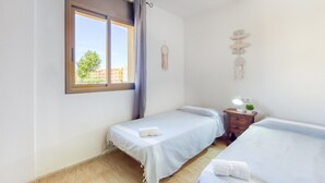 A bedroom with two single beds, a window with a view of buildings, a bedside table with a lamp, and a wall decoration. - Modern Flat by the Sea (Castello d'Empuries)
