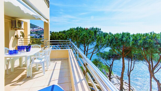 A balcony with a table and chairs, overlooking a coastal area with trees and a view of the sea. - Apartment in Roses Near Sand Beach (Roses)