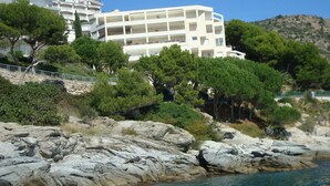 A coastal hotel with multiple floors, surrounded by rocky terrain and dense greenery. - Apartment in Roses Near Sand Beach (Roses)