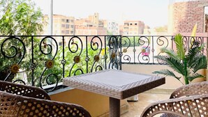A balcony with a table and two wicker chairs, a metal railing with sunflowers, and a view of buildings in the background. - Kemt pyrimds view  (Giza)