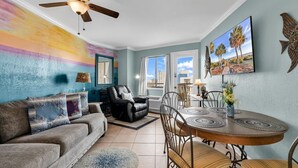 A living room with a sofa, a dining table with chairs, a ceiling fan, and a large window with a view of palm trees and buildings. - Bluewater Resort 1205 (Myrtle Beach)