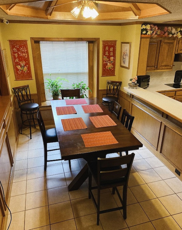 A dining area with a long table, chairs, a kitchen area, and a ceiling fan.