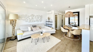 A modern living room with a white sofa, a dining area with wicker chairs, and a kitchen visible through an open door. - Apartment Near Canyelles Petites Beach (Roses)
