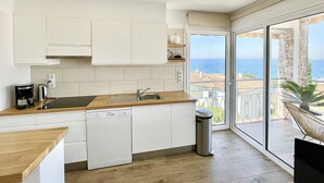 A modern kitchen with white cabinets, a wooden countertop, a dishwasher, and a sink. There is a trash can and a view of the sea through large glass doors. - Apartment Near Canyelles Petites Beach (Roses)