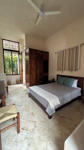 Une chambre spacieuse avec un grand lit, un ventilateur de plafond, une armoire en bois et une vue sur la verdure par la fenêtre.