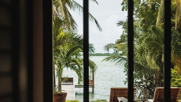 A view through a glass door to an outdoor seating area with a pool, palm trees, and a view of the water. - Ik Casa Del Viento  (Chetumal)