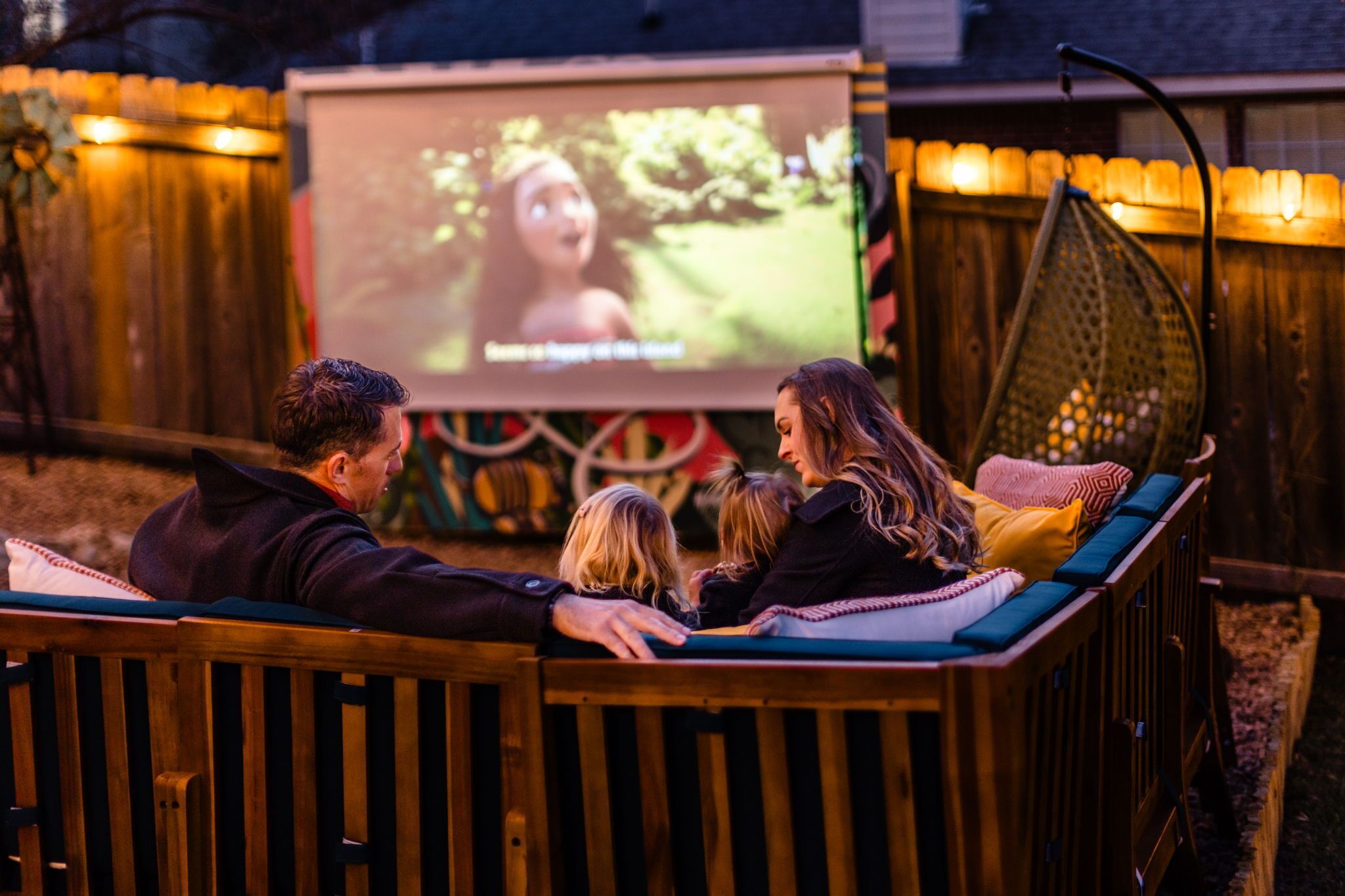 Une famille regarde un film sur un grand écran, installée dans un cadre extérieur agréable, avec un banc en bois et un fauteuil suspendu.