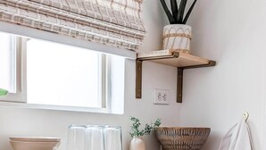 Een keuken met een houten aanrecht, een witte spoelbak en een raam met jaloezieën. Er staan planken met decoratieve objecten en een handdoek hangt aan een haak.