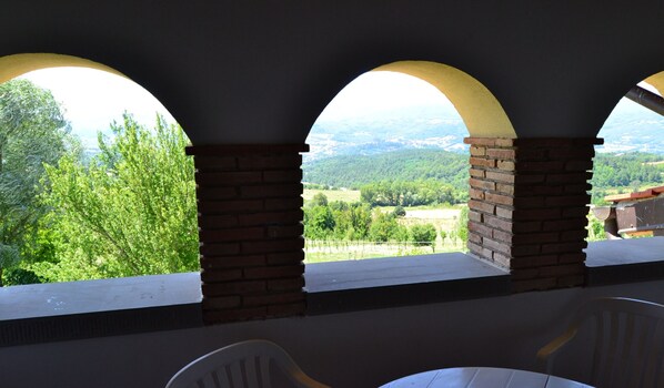 A covered outdoor seating area with brick pillars and white railings, overlooking a green landscape with trees and distant hills. - Farmhouse in Tuscany With Pool and Valley Views (Poppi)