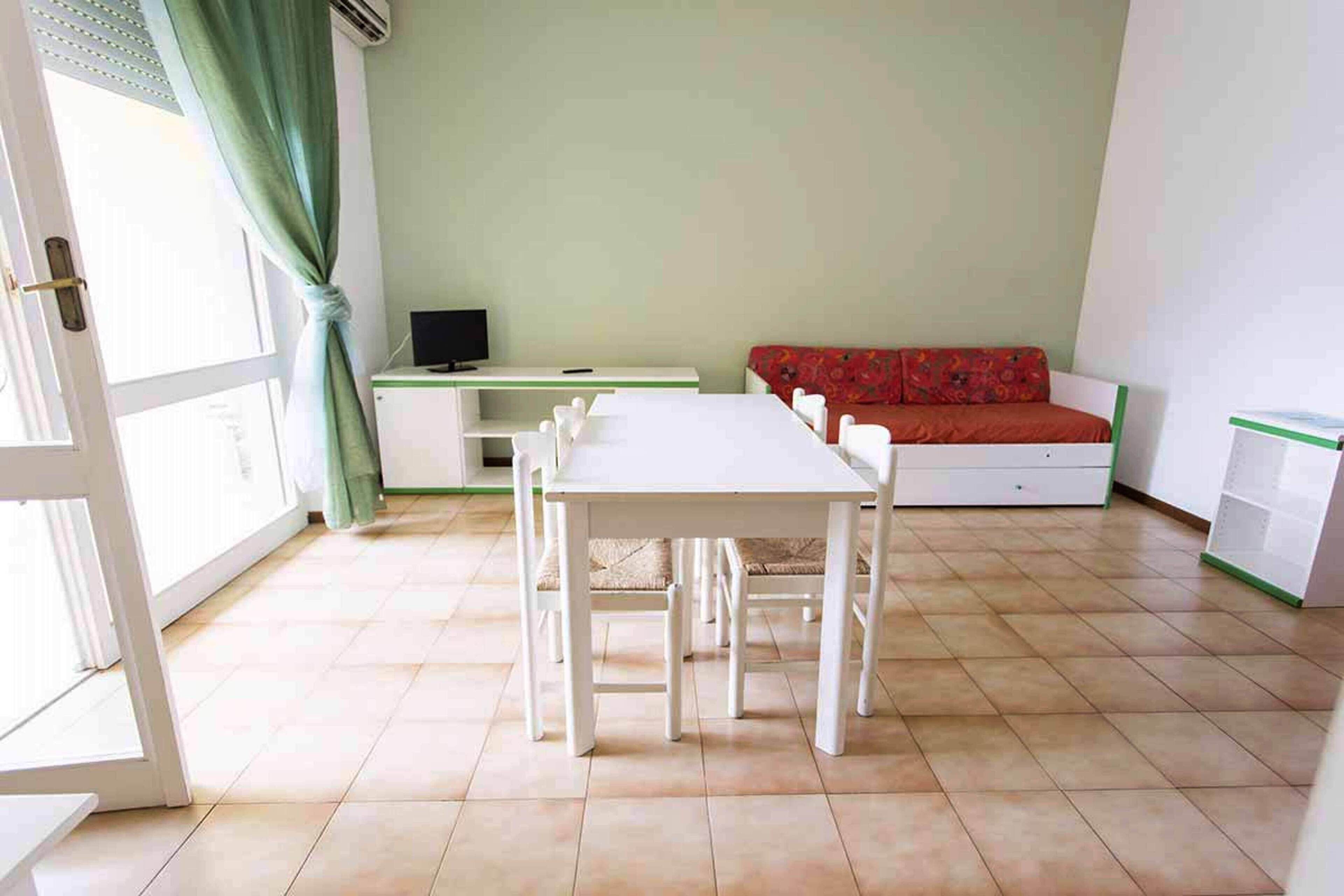 Una habitación con una mesa y sillas blancas, un sofá con cojines rojos, un televisor sobre un mueble blanco y una ventana con cortinas.