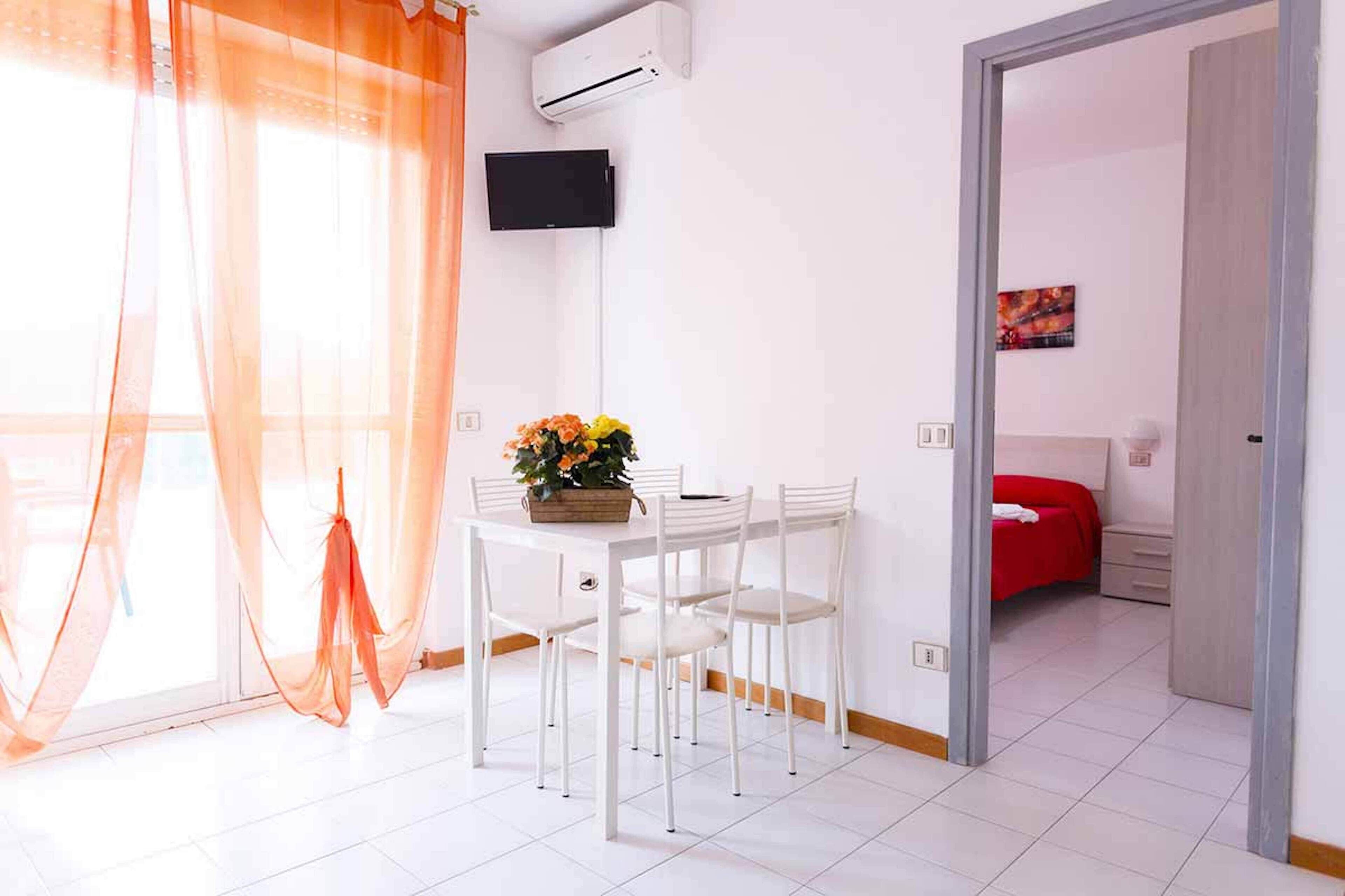 A room with a dining table, chairs, a bouquet of flowers, an air conditioning unit, and a bed with a red cover.