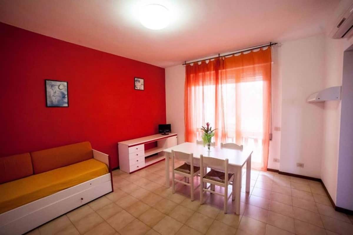 Ein kompakter Wohnraum mit roter Wand, einer Couch, einem Esstisch mit Stühlen, einem kleinen Schreibtisch und einem Fenster mit orangefarbenen Vorhängen.