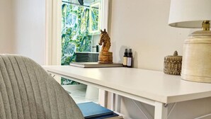 Een kamer met een wit bureau, een grijze stoel, een ingelijst schilderij aan de muur en een raam met uitzicht op groen.