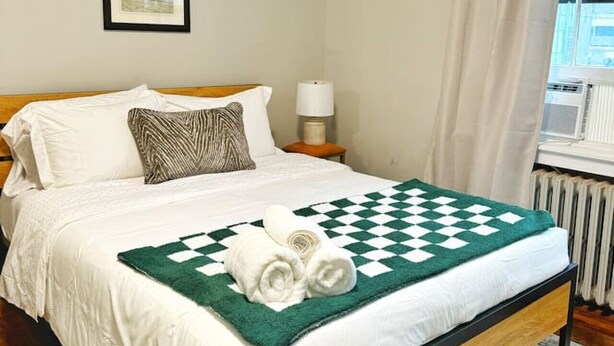 Een netjes opgemaakt bed met een geruit plaid, witte kussens en een ingelijst foto aan de muur.