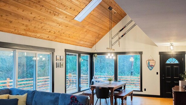 Amplia sala de estar con techo alto, vigas de madera, un sofá grande, una mesa de comedor y ventanas amplias con vistas al exterior.