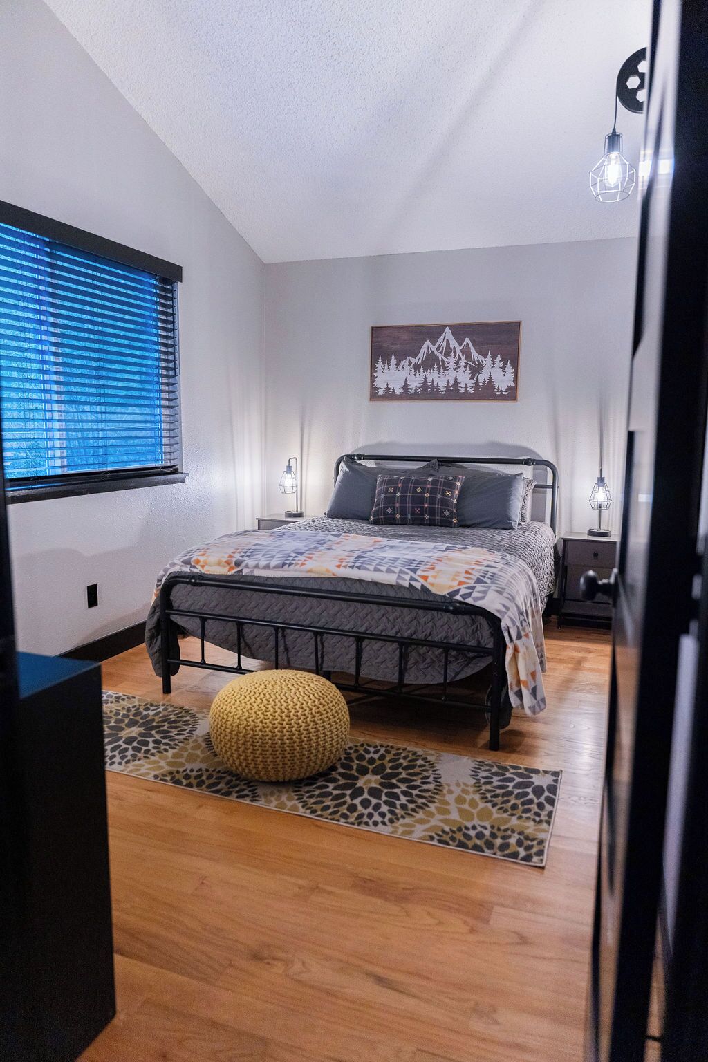 A bedroom with a bed, a window with blinds, a wall-mounted artwork, a bedside table, and a lamp.