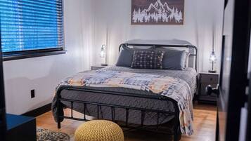 A bedroom with a bed, a window with blinds, a wall-mounted artwork, a bedside table, and a lamp.