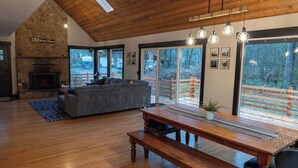 Amplia sala de estar con chimenea de piedra, un sofá grande, una mesa de comedor de madera y ventanas grandes que ofrecen vistas al exterior.