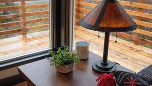 Una mesa de madera con una lámpara, una planta en maceta y una taza junto a una ventana que da a una terraza de madera y árboles.