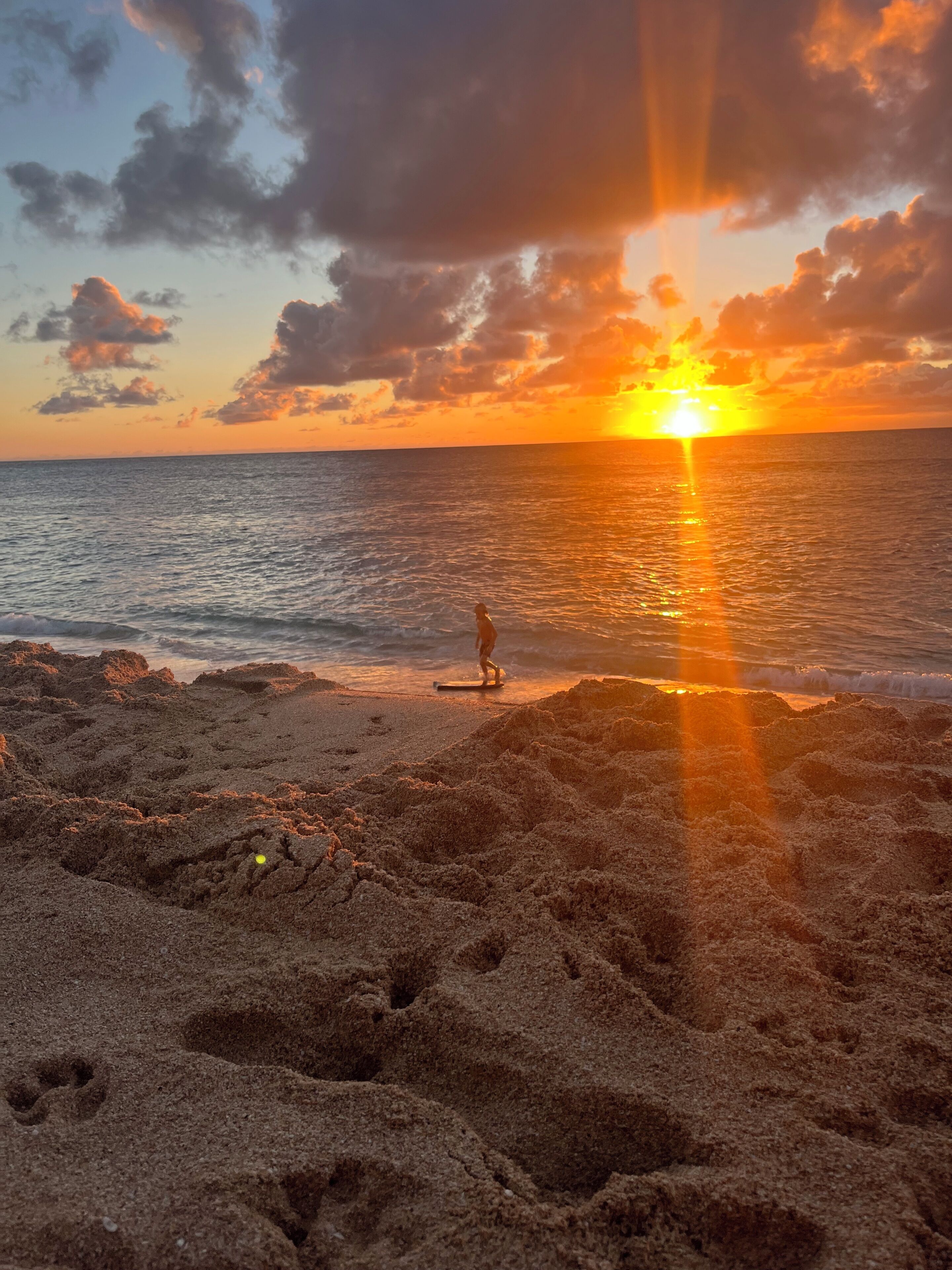 Escena de playa con atardecer, arena y una persona de pie sobre una tabla.