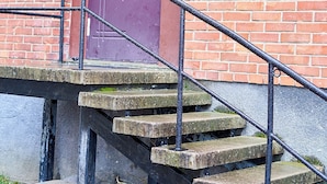 A set of concrete stairs leading to a purple door with a metal handrail, a security camera above, and a utility box on the brick wall. - Tokyo Vibes Hostel with more privacy (Self Check in) (Vilnius)