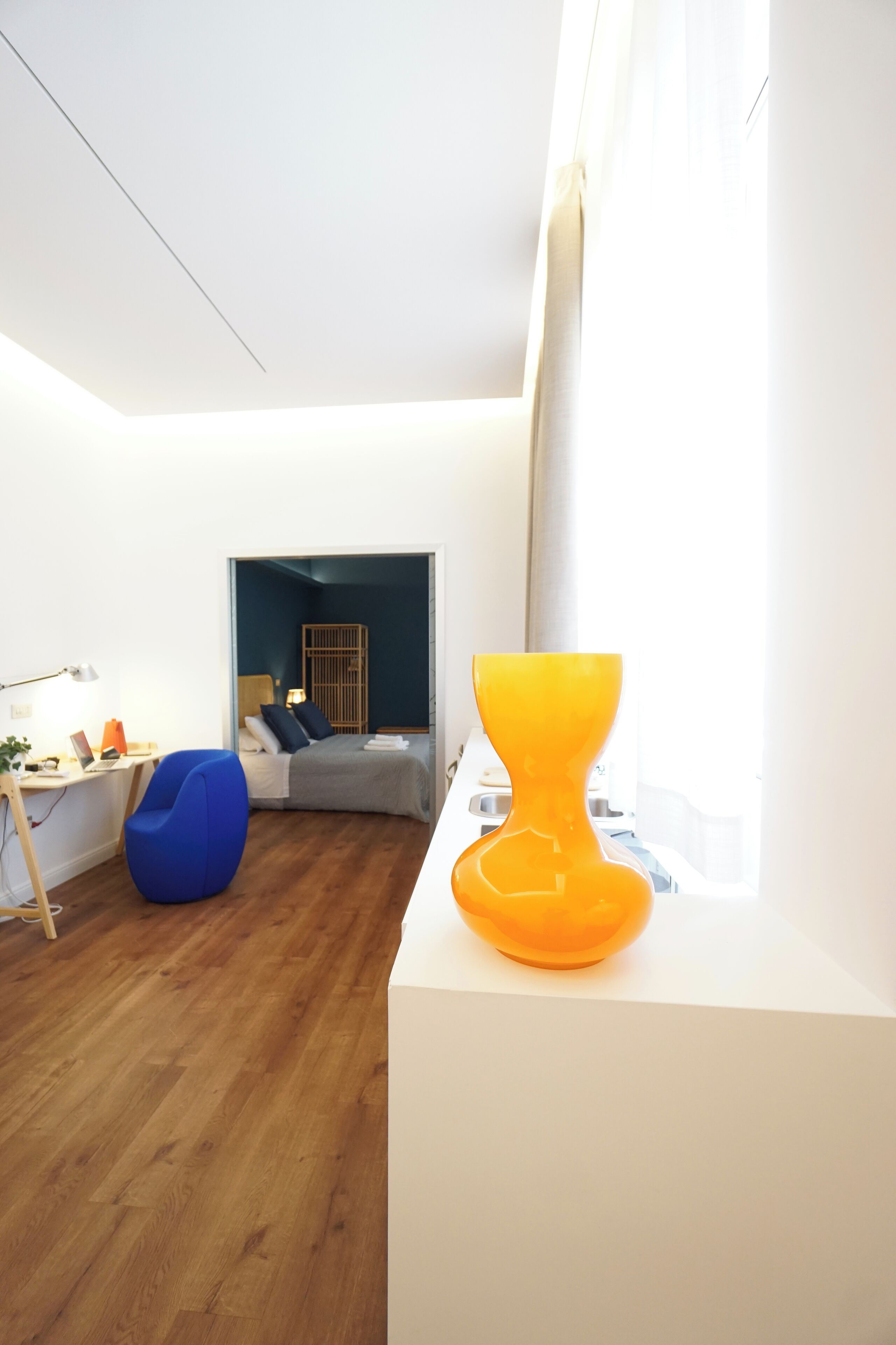 Une pièce moderne et lumineuse, dotée d’un plancher en bois, d’une chaise bleue, d’un lit avec une tête de lit sombre et d’une grande fenêtre.