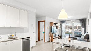 Una cocina moderna con armarios blancos, lavavajillas y un comedor con mesa y sillas de vidrio.