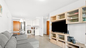 Una sala moderna con un sofá seccional gris, un televisor de pantalla plana sobre un mueble de madera, y una cocina al fondo.