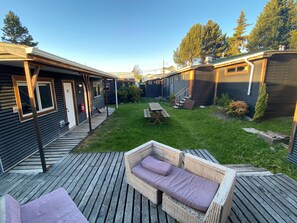A deck with wicker furniture and a picnic table, surrounded by a grassy yard and wooden buildings. - HOSTAL AMERINDIA (Natales)