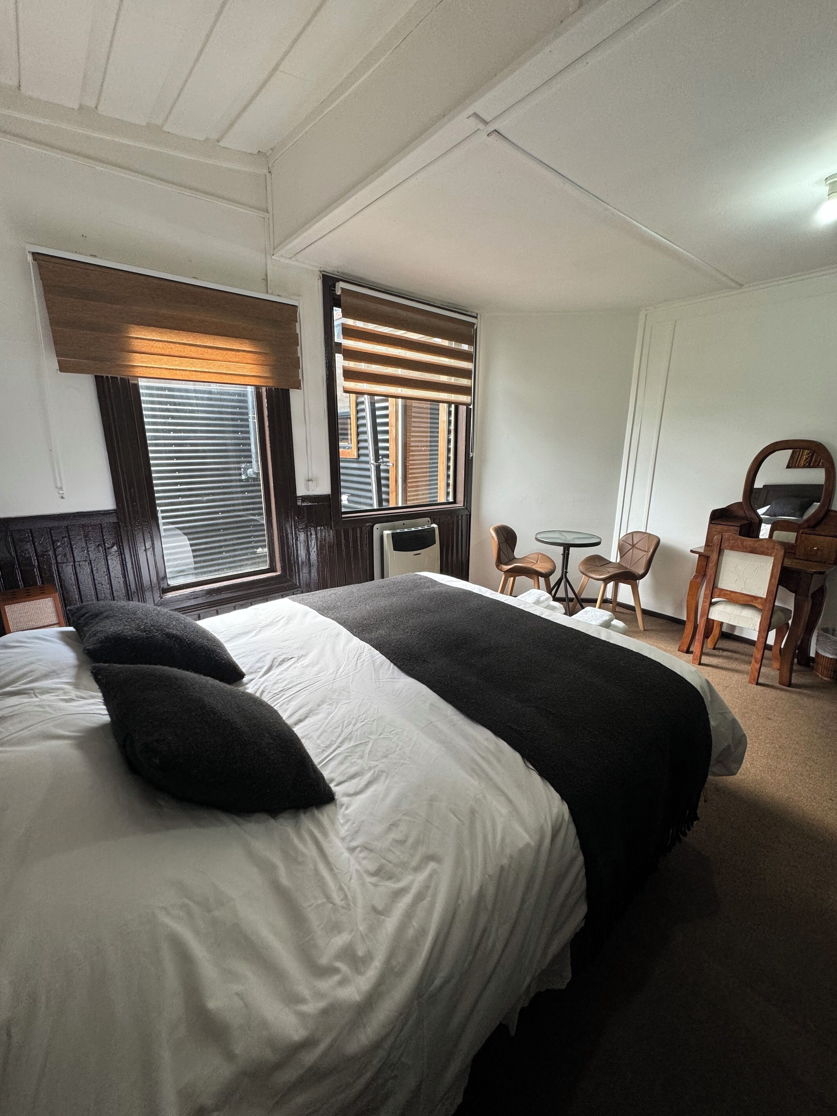 Habitación de hotel con una cama grande, una mesita, dos sillas, un espejo y una ventana con persianas.