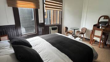 Habitación de hotel con una cama grande, una mesita, dos sillas, un espejo y una ventana con persianas.