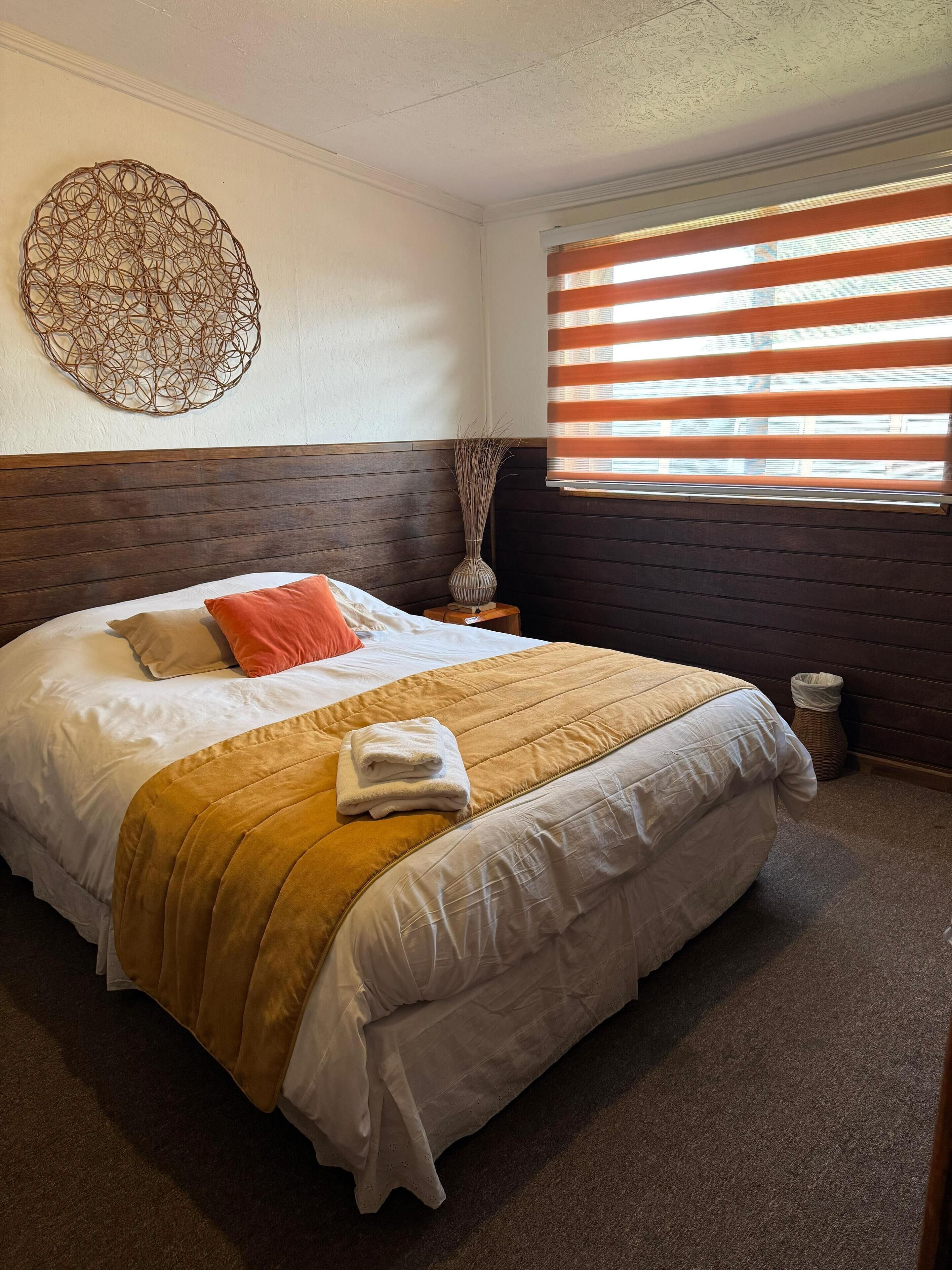 A neatly made bed with a headboard, a bedside table with a vase, and a window with blinds.