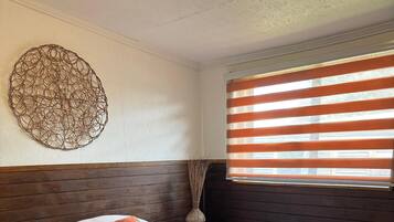 A neatly made bed with a headboard, a bedside table with a vase, and a window with blinds.