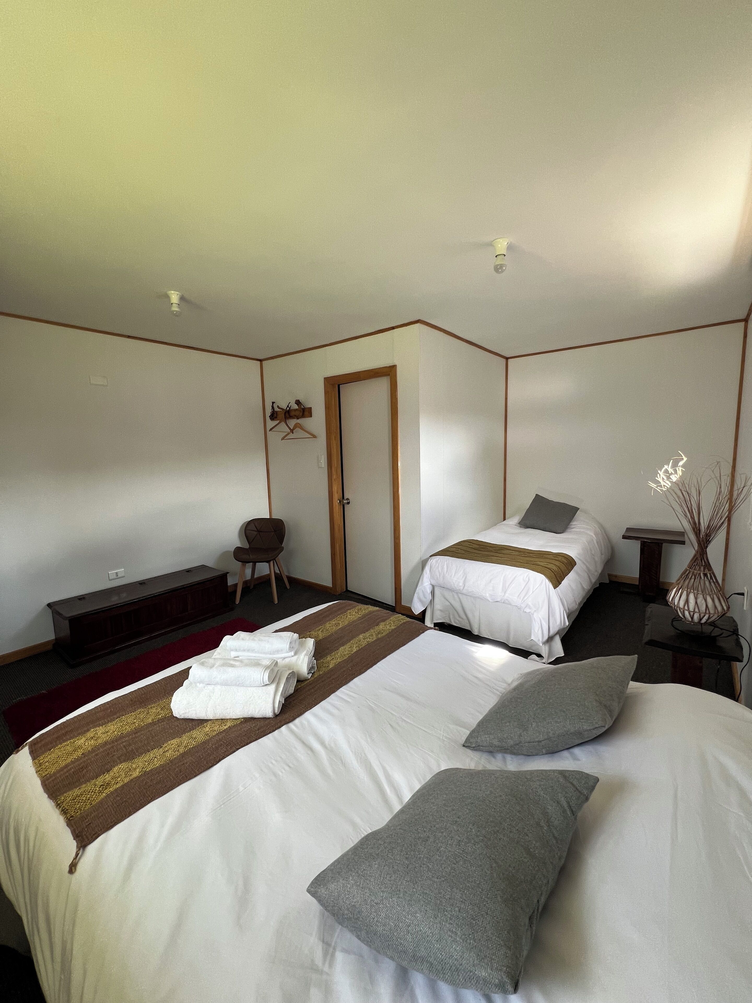 A hotel room with two beds, a bench, a chair, a bedside table, and a wall-mounted shelf.