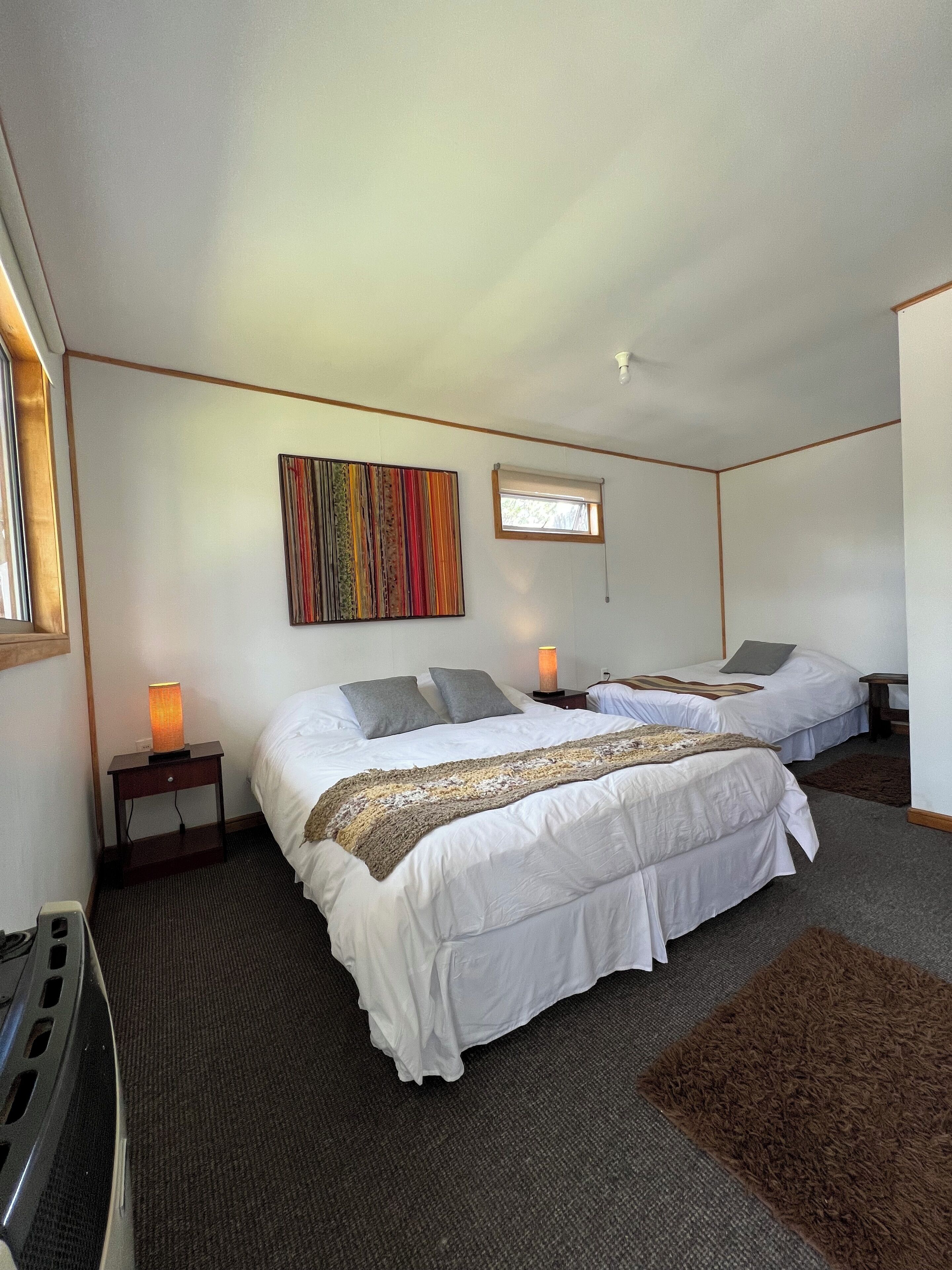 A hotel room with two beds, a colorful artwork on the wall, a window, and a small table with a lamp.
